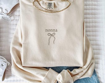 May include: Ivory-colored sweatshirt with the word "nonna" and a bow graphic. The long-sleeved crewneck shirt is folded, with a gold chain necklace draped across it. The text "IVORY SWEATSHIRT" is at the bottom.