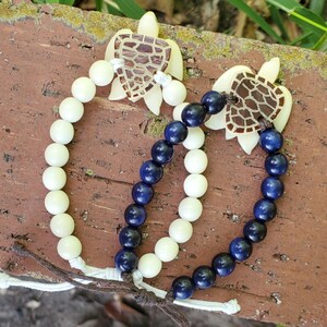 May include: Two beaded bracelets with turtle charms. One bracelet features cream-colored beads, while the other has navy blue beads. The turtle charms are light brown with a cutout design. The bracelets are displayed on a textured, brown surface.