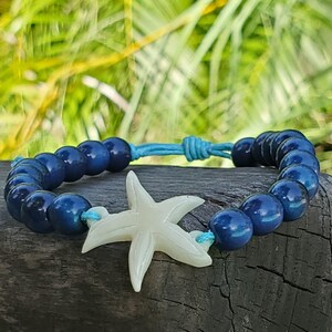 May include: A blue beaded bracelet with a white starfish charm. The bracelet features dark blue, round beads strung on a light blue cord. The starfish charm is a light cream color and is centered on the bracelet.