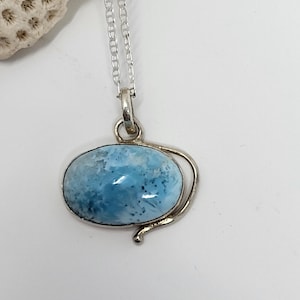 May include: A silver necklace featuring a light blue oval larimar stone pendant. The stone has a marbled appearance with white and darker blue inclusions. The pendant is suspended from a delicate silver chain, creating a simple and elegant design.