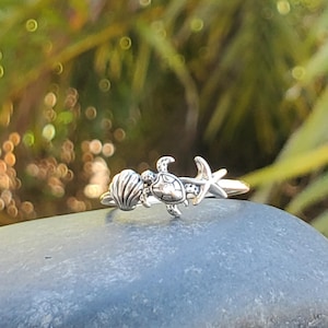 May include: A silver ring with a sea turtle, seashell, and starfish design. The ring is on a gray stone.
