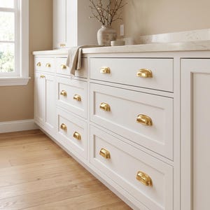 Puede incluir: Un juego de tiradores de cajón tipo concha en latón pulido instalados en armarios blancos de estilo shaker. Cada tirador presenta un diseño liso, redondeado y semicircular con un acabado dorado metálico para abrir cajones y puertas.