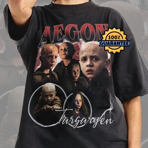 以下が含まれることがあります： 黒いTシャツ。赤い文字で「AEGON」と「100％保証」のバッジが付いた、エゴン・ターガリエンのグラフィック。エゴンと他のキャラクターの肖像画が含まれています。ヴィンテージスタイルのTシャツ。
