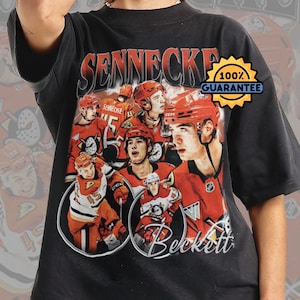 Puede incluir: Camiseta negra con un diseño vintage que presenta jugadores de hockey sobre hielo en rojo y blanco. La camiseta muestra los nombres "Sennecke" y "Beckett". También es visible un sello dorado con "100% Garantía".