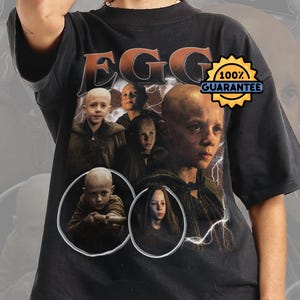 以下が含まれることがあります： オレンジ色の「EGG」グラフィックと、頭を剃った子供たちの画像が特徴の黒いTシャツ。「100％保証」バッジが含まれています。