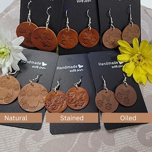 以下が含まれることがあります： 天然、染色、オイル仕上げのハンドメイドレザーイヤリングのコレクション。 各ペアには、フクロウ、蝶、花柄など、ユニークなエンボス加工のデザインが施されています。 イヤリングは、「Handmade with love」のテキストが入った黒いカードに表示されています。