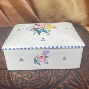 May include: A white porcelain trinket box with a blue and white trim and floral designs in pink, yellow, and green.