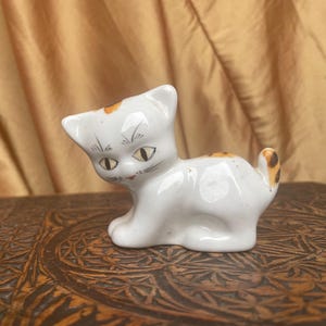 May include: A white porcelain figurine of a cat with orange and brown markings. The cat has a painted face with black whiskers and yellow eyes.