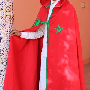 May include: A red hooded cloak with green trim and a green star emblem. The cloak is worn over a white shirt and pants. The outfit is a traditional garment, likely for cultural or ceremonial purposes.