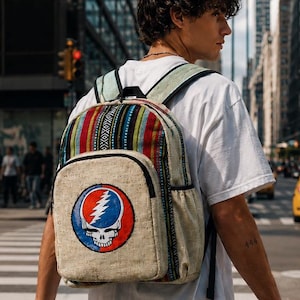 May include: A tan backpack with a colorful striped top section and a Steal Your Face logo on the front. The backpack has adjustable straps and is being worn by a person crossing a city street.