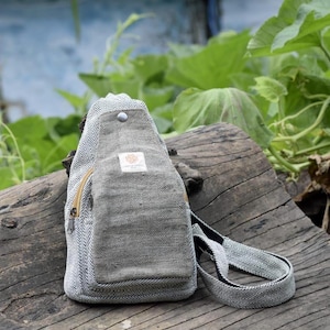 May include: A gray and white herringbone pattern sling bag with a zippered pocket and adjustable strap. The bag is made of a textured fabric and features a small label on the front. The bag is sitting on a weathered wooden surface.