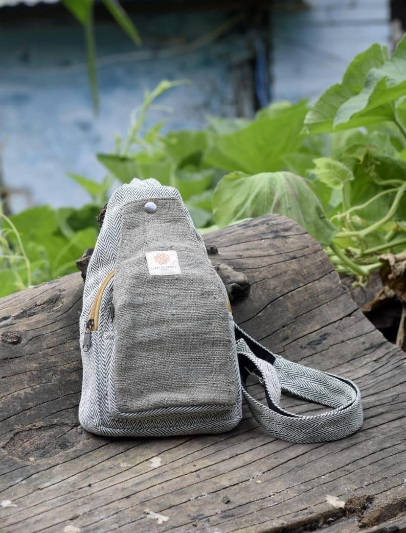 May include: A gray and white herringbone pattern sling bag with a zippered pocket and adjustable strap. The bag is made of a textured fabric and features a small label on the front. The bag is sitting on a weathered wooden surface.