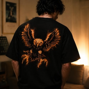 Könnte beinhalten: Ein schwarzes Kurzarm-T-Shirt mit einer großen Grafik auf der Rückseite, die einen Adler mit ausgebreiteten Flügeln zeigt. Der Adler ist in Orange- und Gelbtönen gehalten, was einen leuchtenden, feurigen Effekt auf dem dunklen Stoff erzeugt.