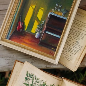 May include: An oil painting in a gold frame depicts a room with a piano, a red guitar, and a vase of flowers. The painting has a warm color palette with sunlight streaming through a window. Old books are in the foreground.