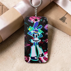 Puede incluir: Un llavero rectangular con un personaje de dibujos animados con cabeza azul, vestido blanco y piernas largas blancas. El fondo es negro con detalles en rosa y verde. El llavero tiene un anillo plateado.