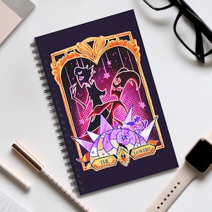 May include: Spiral-bound notebook with a dark purple cover featuring a colorful illustration. The artwork includes two stylized figures within a gold-framed heart, with the words "THE LOVERS" on a banner. The design has a fantasy theme with pink and purple accents.