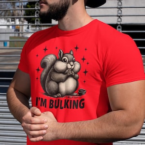 Könnte beinhalten: Rotes T-Shirt mit einem Cartoon-Eichhörnchen und dem Text "I'M BULKING". Das Eichhörnchen ist grau und weiß, mit einem großen Bauch und einer kleinen Schüssel. Schwarze Sterne umgeben das Eichhörnchen.