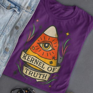May include: A purple t-shirt with a candy corn graphic, an eye, and the words "KERNEL OF TRUTH". The image includes a denim jacket, sunglasses, and beaded bracelets. The design is eye-catching and unique.