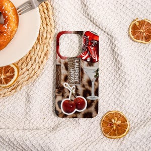 May include: A phone case with a leopard print background and various stickers. Stickers include two Coca-Cola cans, a martini glass, two cherries, and text that reads "LAURENT SAINTS".