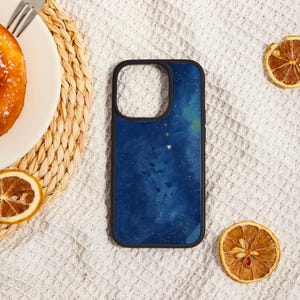 May include: A smartphone case with a dark blue watercolor design featuring gold stars and a black border. The case is lying on a white surface with dried orange slices and a plate with a pastry.