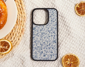 May include: A phone case with a black border and a light blue floral pattern. The case is lying on a white textured surface, with a plate of food and dried orange slices nearby.