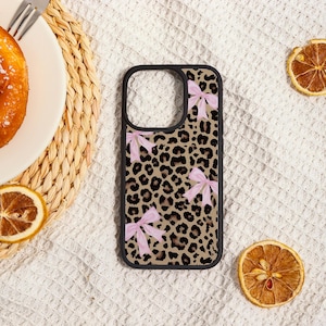 May include: A black phone case with a leopard print design and pink bow accents. The case has a black border and is set against a white textured background with dried orange slices.