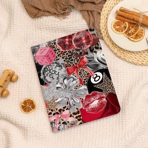 May include: A decorative tablet case featuring a collage of playful elements. The design includes a pink dice with the number 7, disco balls, red lips, a silver flower, and a leopard print background. A red bow and the number 8 ball are also visible.