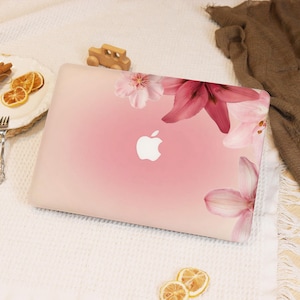 May include: A pink laptop case featuring a floral design with pink and white lilies. The Apple logo is visible in the center. The case is on a white surface with a wooden toy car and a plate of dried citrus slices.