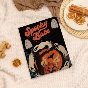May include: Black notebook with the text "Spooky Babe" in orange script. The cover features a Halloween-themed illustration with a jack-o'-lantern, ghosts, and a bat silhouette against a full moon. The design has a vintage, retro aesthetic.