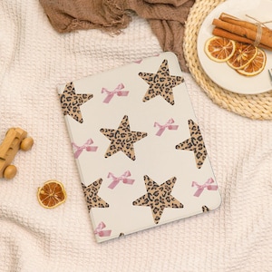 May include: A cream tablet case with a pattern of leopard print stars and pink bows. The case is rectangular with rounded corners. The background is a white, textured surface.