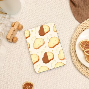 May include: A rectangular notebook featuring a repeating pattern of bread slices, some dipped in chocolate, on a cream background. Gold star accents and radiating lines surround the bread. A wooden toy car and other items are in the background.