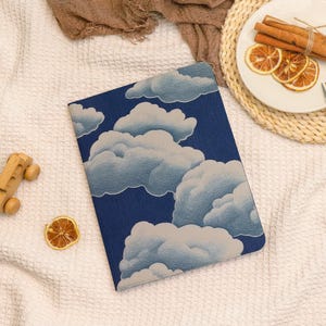 May include: A blue notebook with a cloud design. The cover features a dark blue background with white and light blue clouds. The notebook is lying on a white textured surface, with a wooden toy car and dried orange slice nearby.
