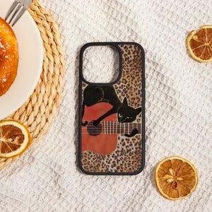 May include: Black phone case with a leopard print background and a black cat playing a red guitar. The case has a black border and is on a white surface with a plate of food and dried orange slices.