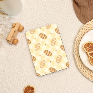 May include: A rectangular mousepad with a yellow and white gingham pattern, featuring brown bread loaf illustrations. The mousepad is on a white surface, with a wooden toy car and a decorative plate with dried orange slices.