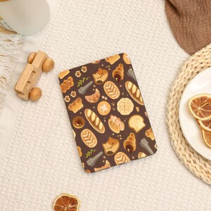 May include: A brown notebook featuring a repeating pattern of various baked goods, including croissants, bread loaves, and pastries. The illustrations are in shades of brown and tan, with a few green sprigs. The notebook is on a white surface.