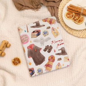 May include: A white tablet case with a watercolor design featuring a wizarding theme. The case is decorated with illustrations of a wizard's hat, scarf, books, and the words "Happy Potter".