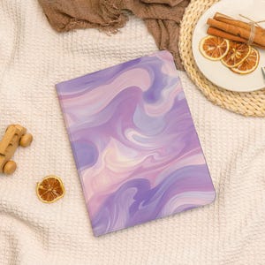 May include: A notebook with a purple, pink, and white swirled design. The notebook is lying on a white textured surface. A wooden toy car and a dried orange slice are also visible.