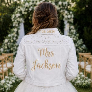 Puede incluir: Una chaqueta vaquera blanca con el cuello bordado con la fecha 25.07.2026 en dorado. La parte trasera está adornada con perlas blancas dispersas por los hombros y el texto Mrs Jackson en purpurina dorada centrado bajo las perlas.