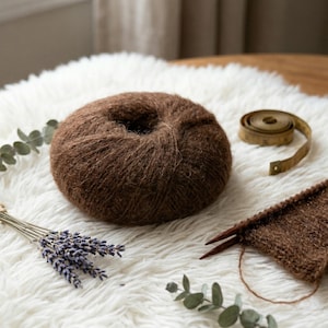 May include: A ball of brown yarn sits on a white, fluffy surface, next to a measuring tape and a small knitting project. Dried lavender and eucalyptus sprigs add a natural touch. The yarn is a rich, warm brown, perfect for knitting or crocheting projects.