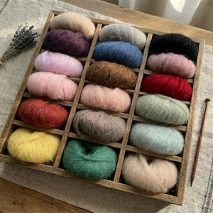 May include: A wooden tray filled with various colored yarn balls. The yarn colors include beige, purple, gray, blue, black, pink, brown, red, green, yellow, and light blue. A wooden knitting needle is placed on the right side of the tray.