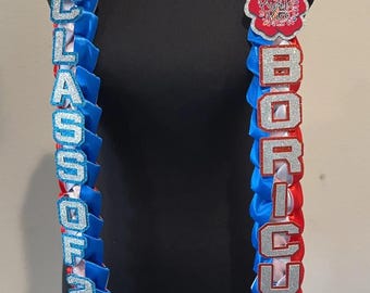 May include: A blue, red, and white lei with the text "Boricua lei" and "Class of 2023". The lei features the Puerto Rican flag and heart-shaped accents. The lei is displayed on a black mannequin.