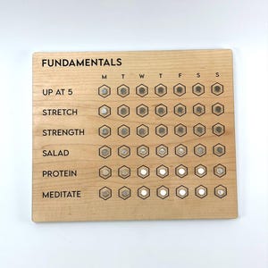 May include: Wooden "Fundamentals" checklist with the text "UP AT 5", "STRETCH", "STRENGTH", "SALAD", "PROTEIN", and "MEDITATE". Each activity has a series of hexagonal spaces for tracking progress. The board is light brown.