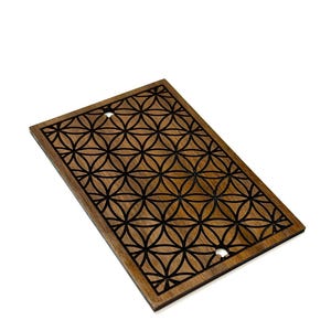 May include: A rectangular wooden panel with a geometric flower of life pattern. The panel is made of dark brown wood with a black design. Two holes are visible near the top. The panel is angled against a white background.