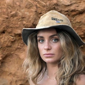 May include: A woman with long blonde hair wearing a brown and grey hat with the text "Afro Blonde" on it. She is wearing a light brown knotted top.