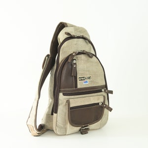 Könnte beinhalten: Beige Canvas-Umhängetasche mit braunen Lederakzenten und mehreren Reißverschlussfächern. Die Tasche trägt den Schriftzug "AFRO BLONDE" auf einem Aufnäher. Sie hat einen gepolsterten Schultergurt und eine kleine Seitentasche.