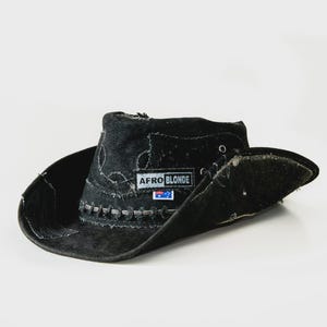 May include: Black cowboy hat with a wide brim and a distressed finish. The hat has a patch that reads "AFRO BLONDE" with a small flag emblem. The hat has a rugged, vintage aesthetic.