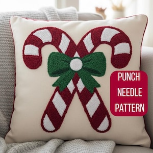 Peut inclure: Coussin crème avec une bordure rouge, orné d'un motif au point de croix de deux cannes en bonbon et d'un nœud vert. Les cannes sont rayées rouge et blanc. Le texte "PUNCH NEEDLE PATTERN" est dans un encadré rouge.