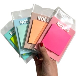 May include: A hand holds a selection of colourful sticky note pads. Each pad is a different colour, including blue, green, orange, and pink. The word "Note" or "Notes" is printed on each pad in white, 3D lettering. The pads are packaged in clear plastic.