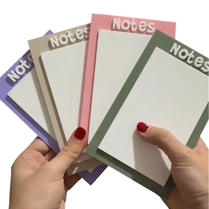 May include: A person's hands holding a stack of memo pads in various colours. Each pad features a white note area, a coloured border, and the word "Notes" in white lettering. The pads are in shades of purple, beige, pink, and green.