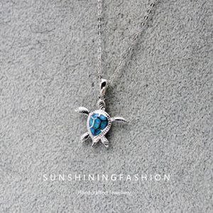 Puede incluir: Un collar de plata con un colgante de tortuga. El colgante tiene un centro azul y negro similar al ópalo y aletas y cabeza detalladas. El collar se muestra sobre un fondo gris. El texto "SUNSHININGFASHION Handcrafted Jewellery" está en la parte inferior.